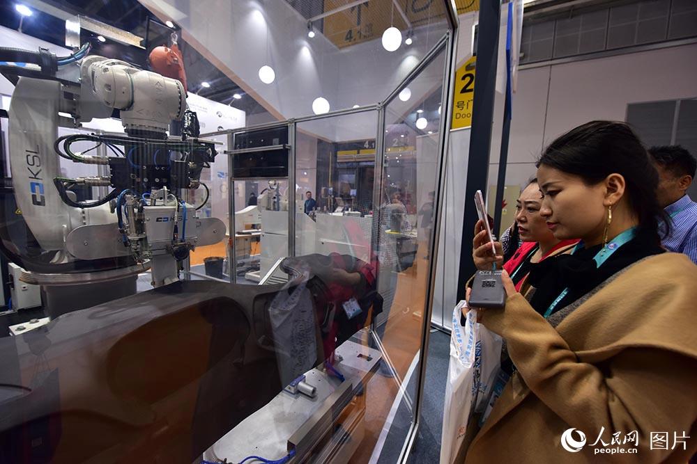 11月5日，在进博会智能及高端装备展区，参观者正在用手机拍摄数字化缝纫机器人。（人民网记者 翁奇羽 摄）