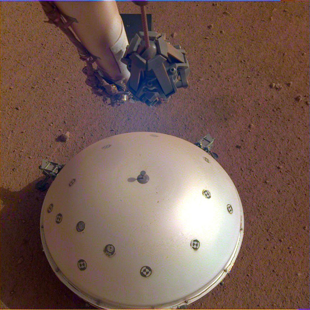 圖為登錄器上一個攝像頭拍攝的照片，展示了火星車的圓形風熱屏障，覆蓋著內部結構地震實驗裝置，背景為火星表面。圖片來源：NASA官網 
