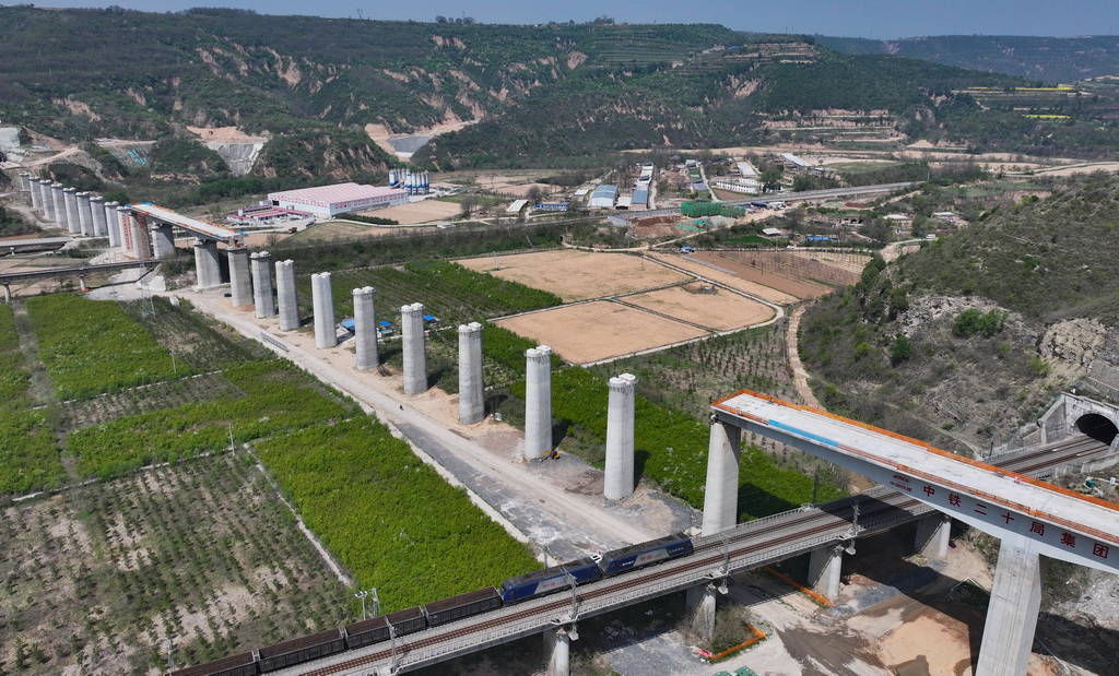 4月17日，在陝西省延安市洛川縣，一列火車從施工中的西延高鐵北洛河特大橋下駛過（無人機照片）。