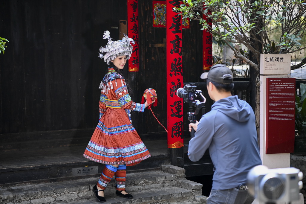 2025年3月23日,在广西南宁市一处少数民族特色建筑展示区内,身穿民族服饰的游客(左)拿着绣球拍摄旅游短视频。新华社记者 曹祎铭 摄